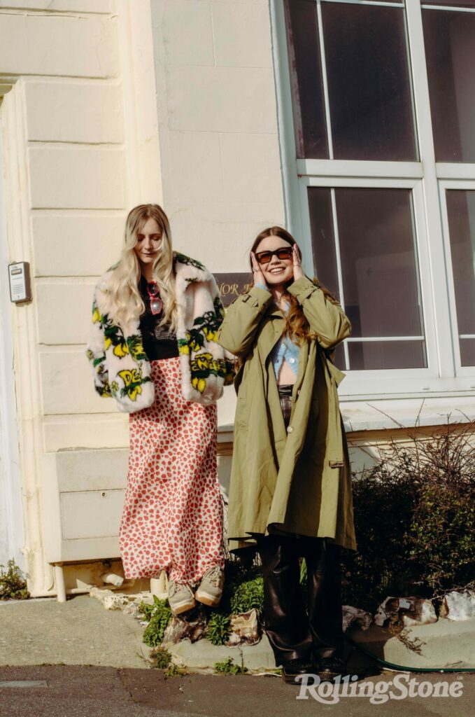 Wet let pose outside a hotel, Rhian wears sunglasses and a long coast, and Hester wears a fluffy jacket and long skirt