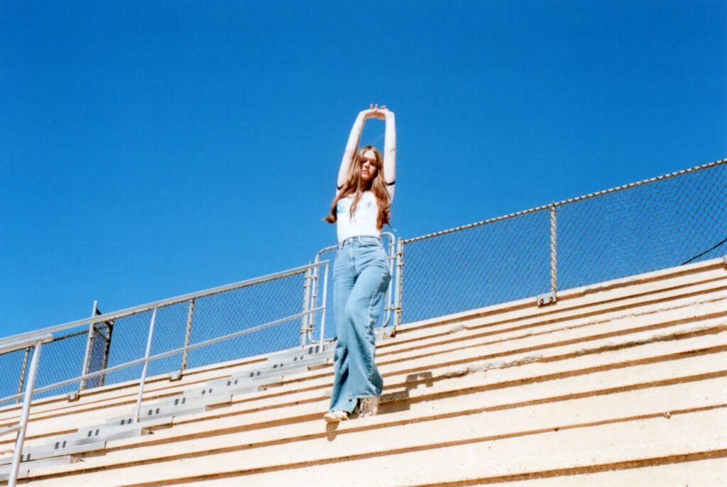 Ethel Cain stands on bleaches with her hands in the air against a blue sky