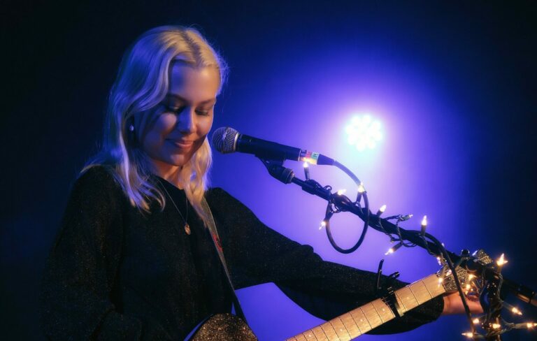 Phoebe Bridgers holds a guitar and stands against a microphone