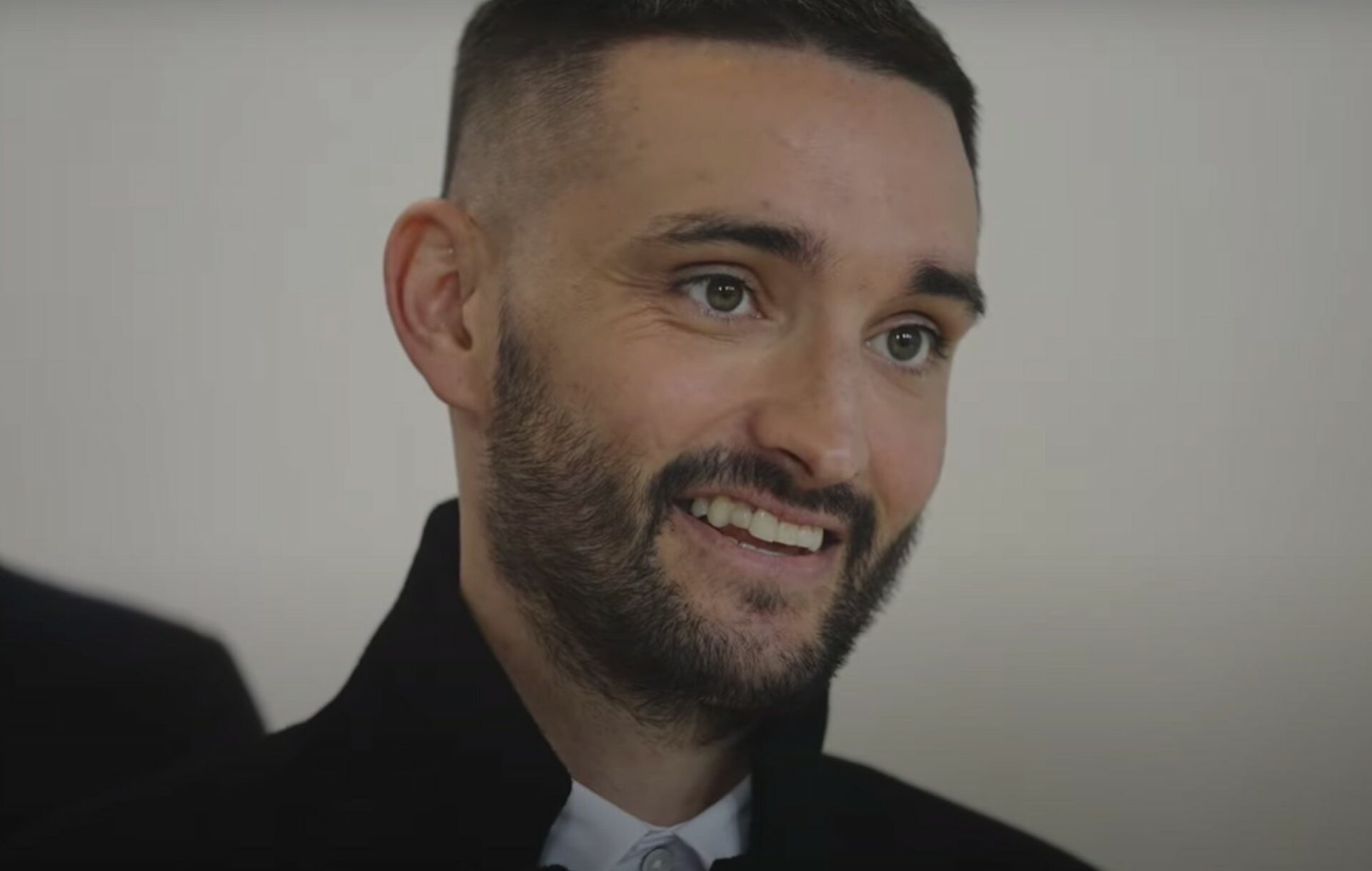 Tom Parker wears a black coat over a button up shirt in a headshot