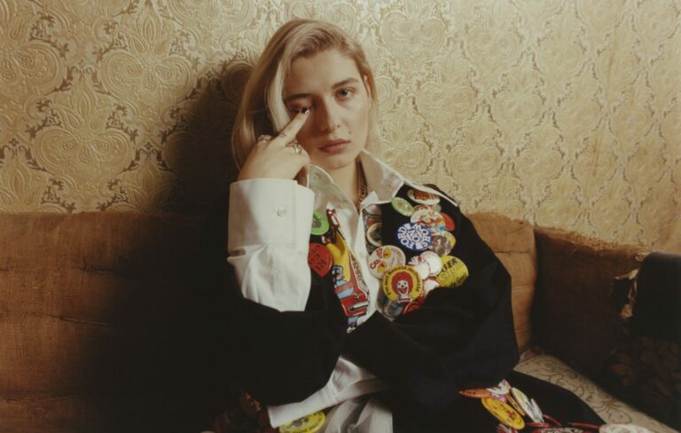 Baby Queen puts her middle finger to her eye, wearing a black jacket covered in badges