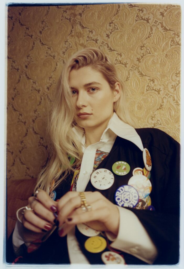 Baby Queen looks to the camera in a white collared shirt and black jacket covered in buttons, her hair is blonde
