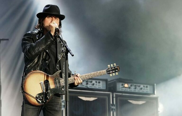 Carl Barat wears a hat, black leather jacket and plays guitar on stage