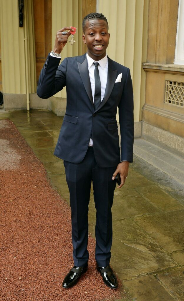 Jamal Edwards wearing a black suit holding up a MBE medal