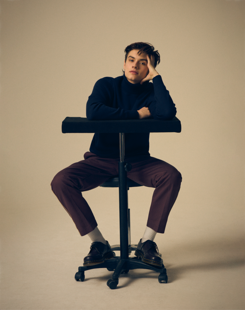 Louis Partridge sits at a desk in dark trousers and a blue shirt