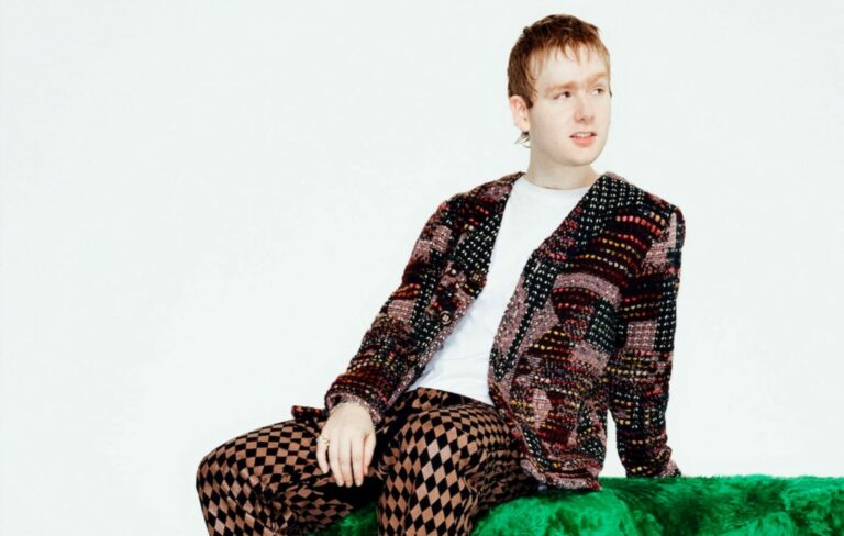 Mura Masa wears a textured suit jacket and checkered trousers on a green chair