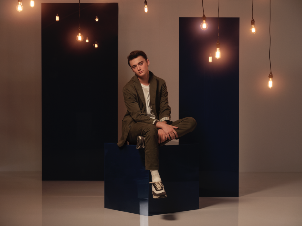 Noah Schnapp wears khaki green and sits on a black box underneath hanging lightbulbs