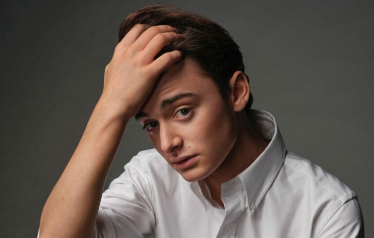 Noah Schnapp puts his fingers in his hair and wears a white collared shirt in a press shot