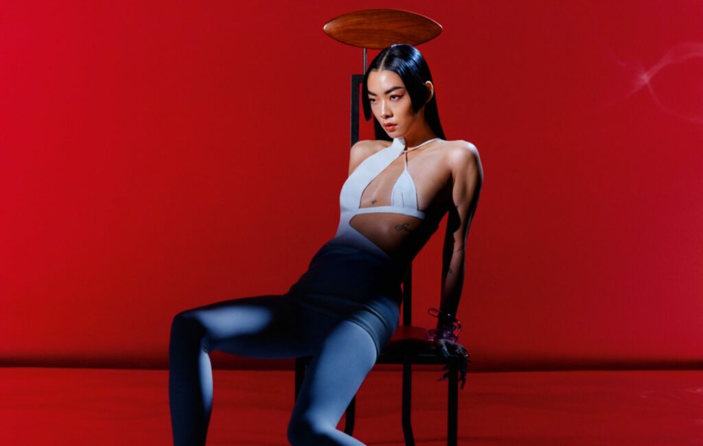 Rina Sawayama wears a white jumpsuit on a chair against a red background