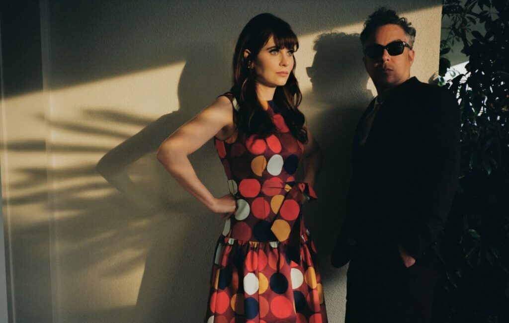 She & Him stand together for a shadowy press shot in a polka dot dress and black suit