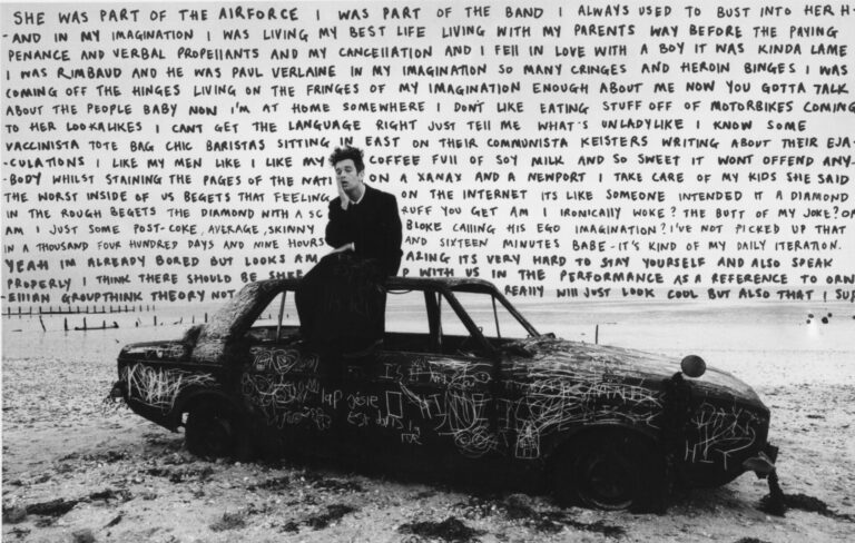 Matty Healy sits against a burnt car with lyrics on a billboard behind him