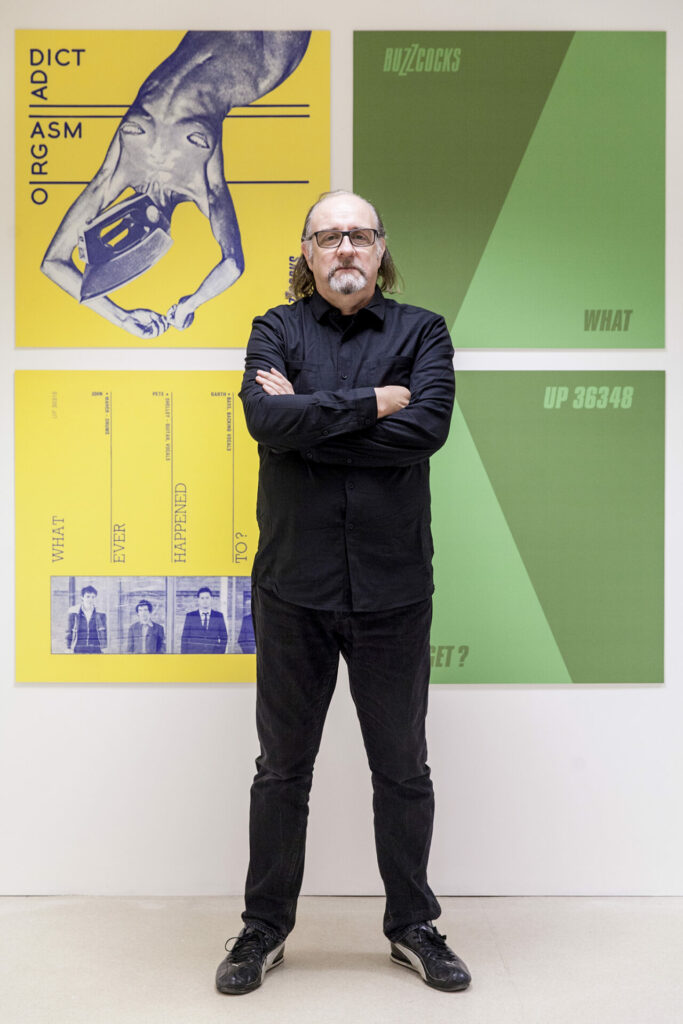 A photograph of artist Malcolm Garrett at an exhibition with his artwork for the Buzzcocks