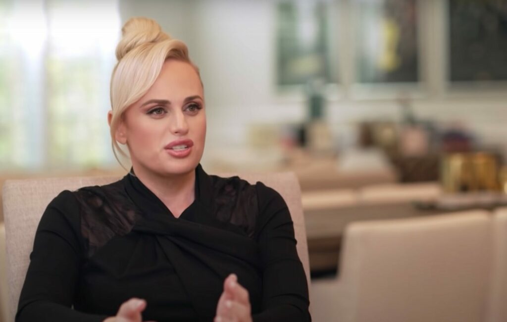 Rebel Wilson wears her hair in a blonde bun and a black shirt in a BBC interview