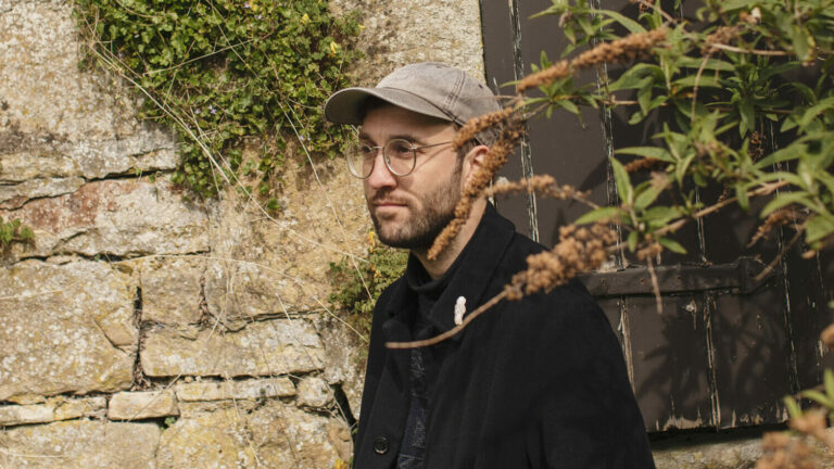 Joe Zadeh stands in front of a stone wall, wearing a black jacket and grey hat