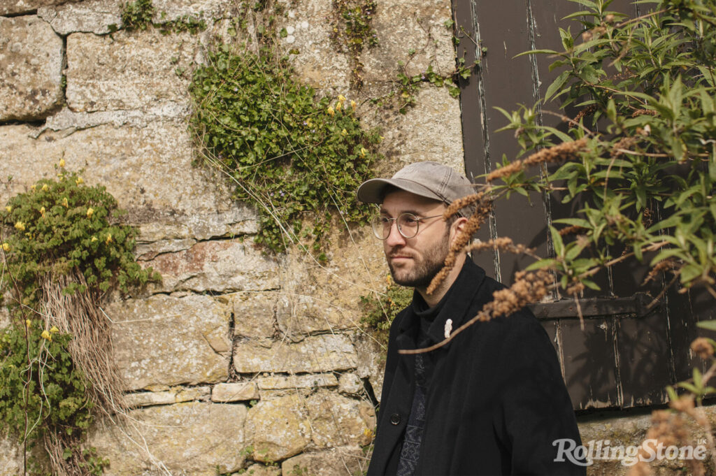 Joe Zadeh stands in front of a stone wall, wearing a black jacket and grey hat