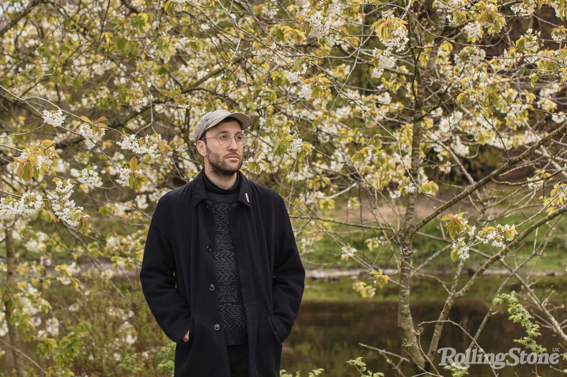 Joe Zadeh stands in front of a tree, wearing a black jacket and grey hat