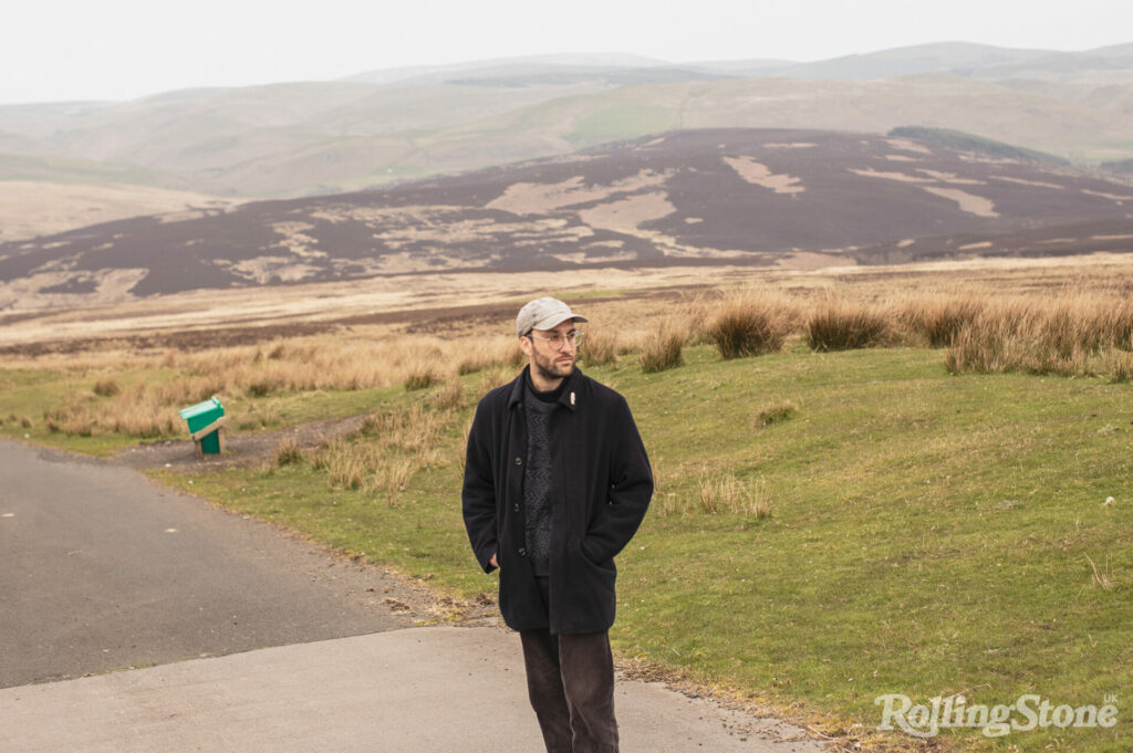 Joe Zadeh stands in front of grassy hills, wearing a black jacket and grey hat
