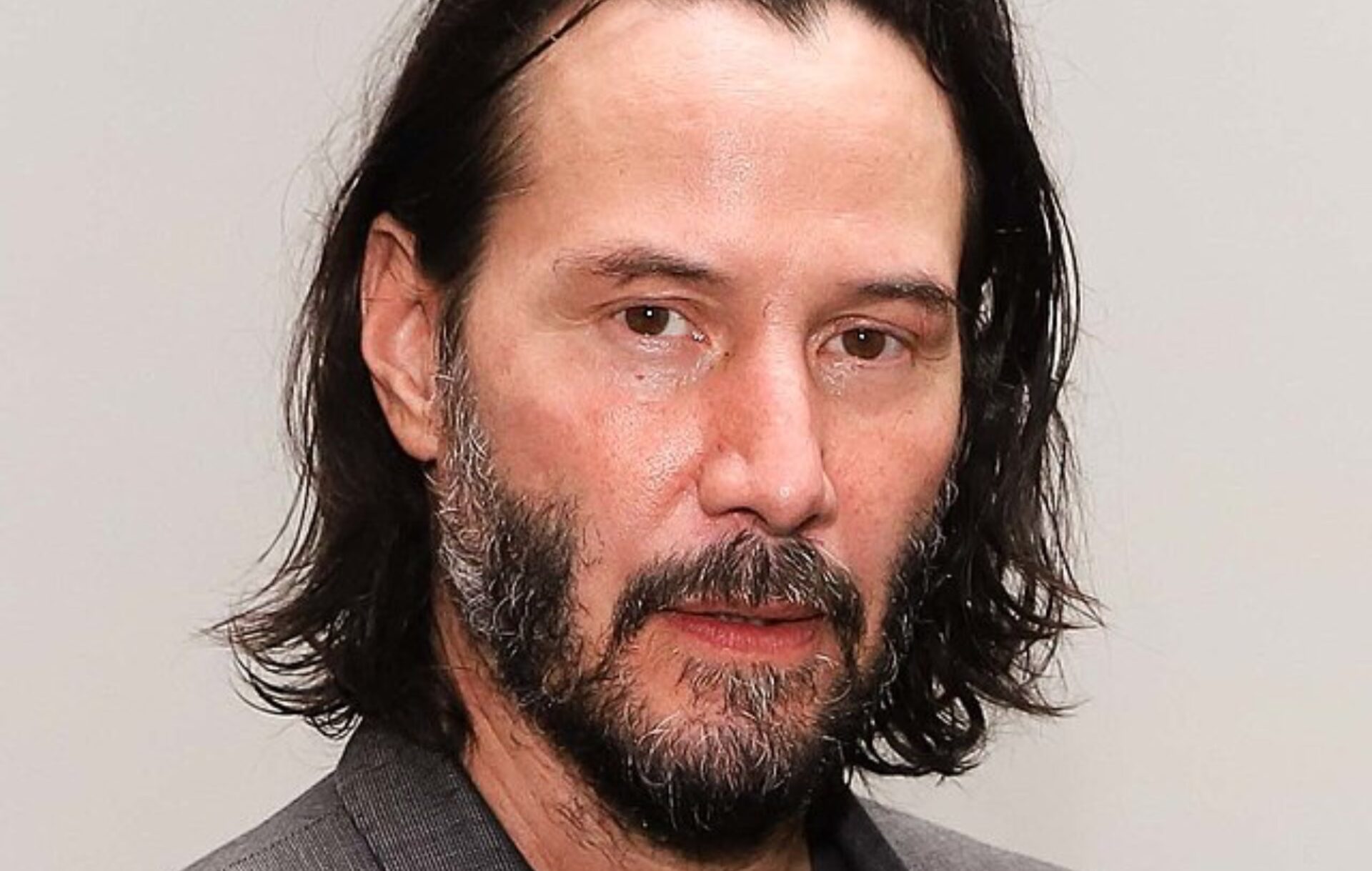Keanu Reeves has long hair and a trimmed beard in a headshot. 