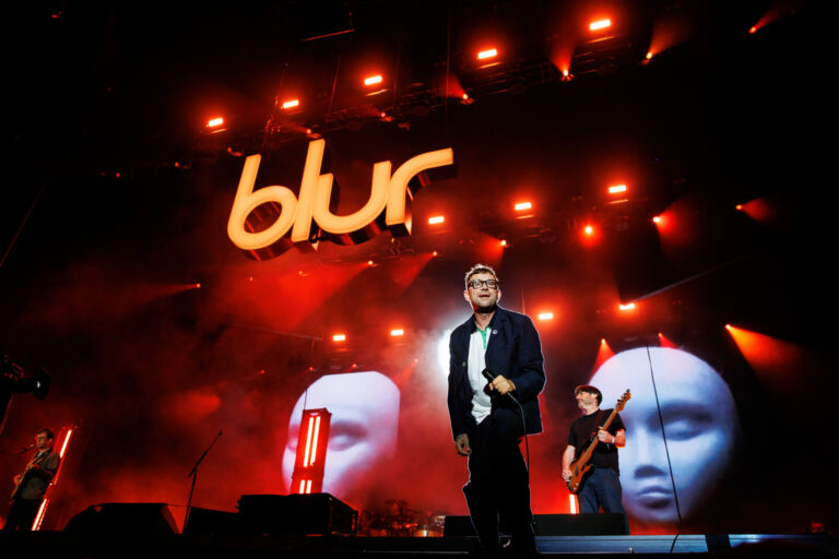 Damon Albarn from Blur stands on a stage with red lights behind him