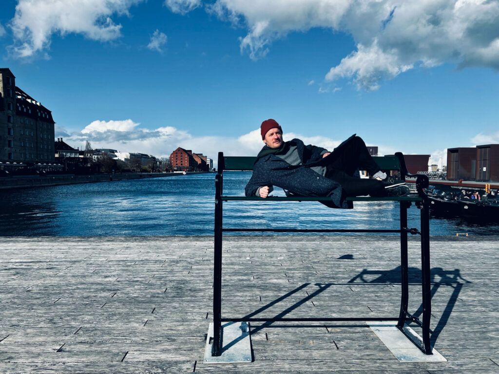 A man lies on a tall bench in Copenhagen against the backdrop of a blue river