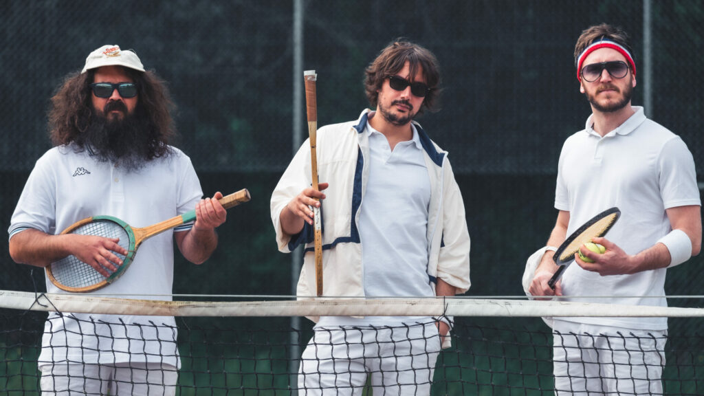 Three men wearing tennis clothes and sunglasses stand behind a tennis net