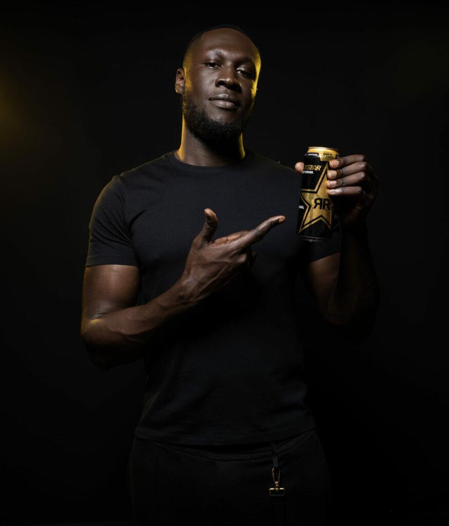Stormzy poses in a black outfit while holding a Rockstar Energy drink can