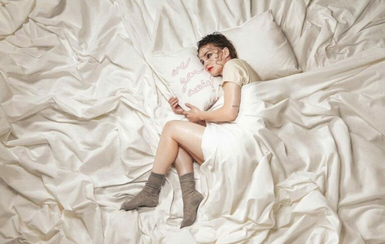 Lauren Mayberry lays on a pile of white bed sheets in a single press shot