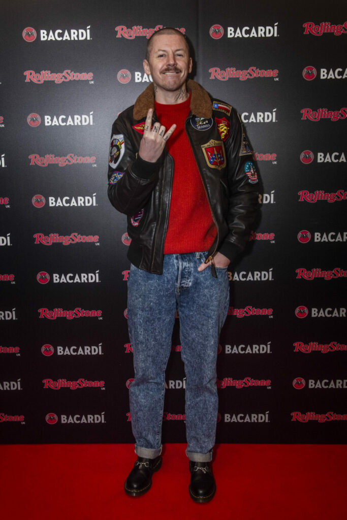 A person stands on a red carpet in front of a step and repeat board with  BACARDÍ and Rolling Stone UK logos