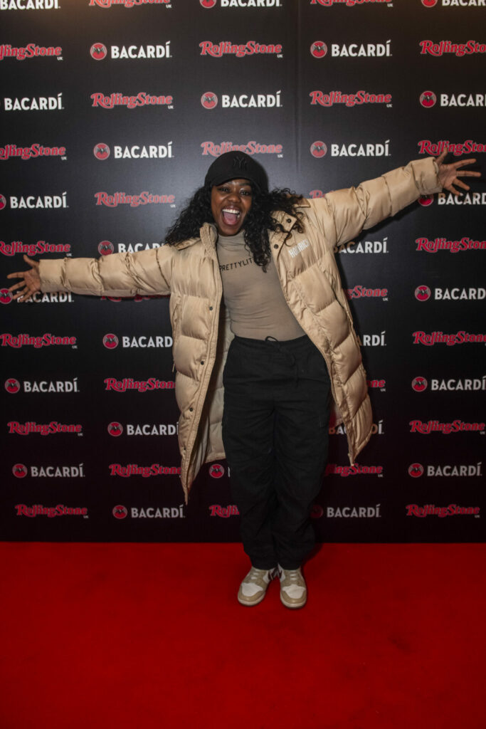 A person stands on a red carpet in front of a step and repeat board with  BACARDÍ and Rolling Stone UK logos