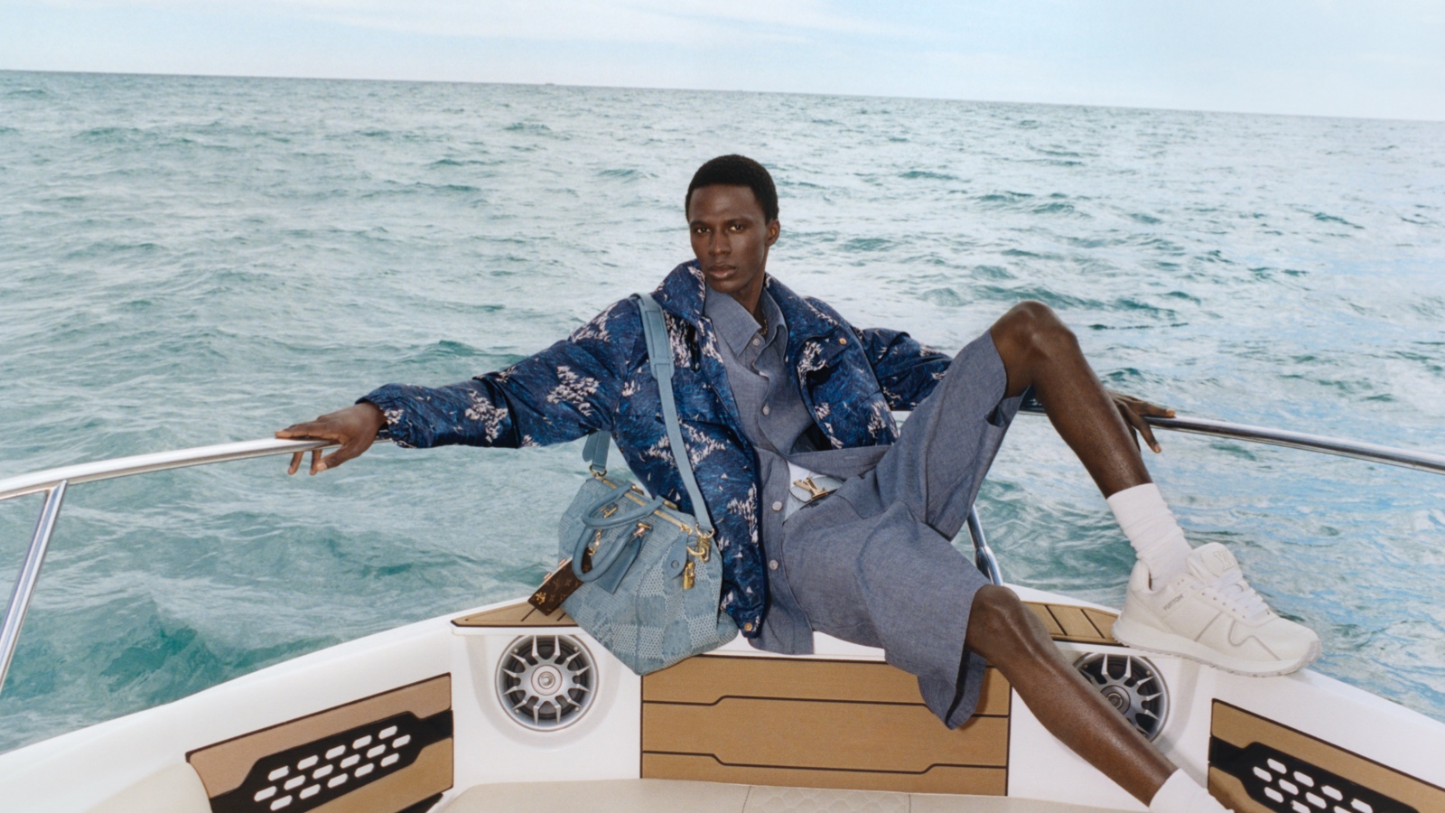 Models poses on edge of boat with monogram blue jacket, bag and plain blue shorts