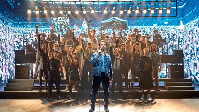 Craige Els as Bob Geldof leads the cast of Just For One Day – The Live Aid Musical in a group performance at The Old Vic, with a backdrop evoking the energy of the iconic 1985 concert.