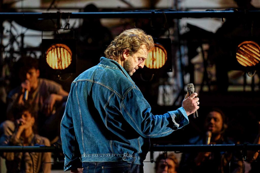Craige Els as Bob Geldof in Just For One Day – The Live Aid Musical, holding a microphone and facing the crowd in a dramatic moment, with stage lights and cast members in the background.