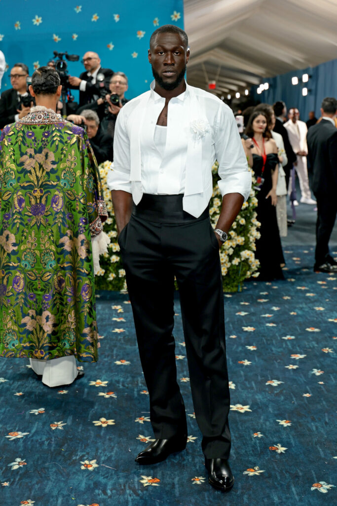 Stormzy wears white ruffled shirt over a white vest, accessorized with a white floral brooch by Haider Ackermann x Tom Ford