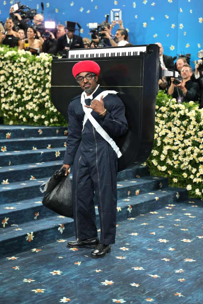 André 3000 wears Burberry boiler suit, red hat with baby grand piano attached to his back