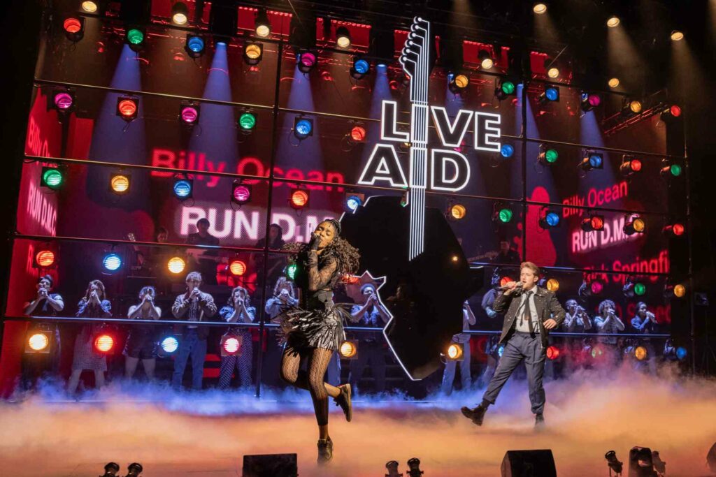 Performers on stage in Just For One Day – The Live Aid Musical, dancing in front of a backdrop featuring the Live Aid logo and names of iconic acts like Billy Ocean and Spandau Ballet.
