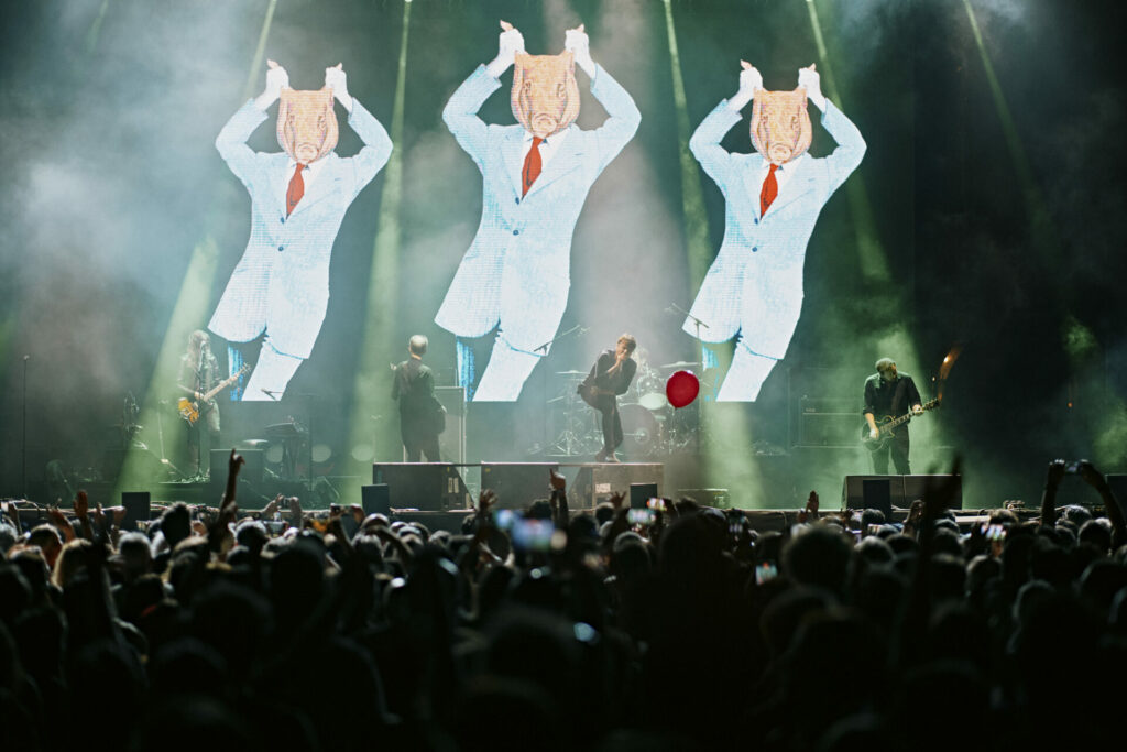 Suede performing at Mallorca Live Festival 2025, with animated visuals of dancing figures in suits displayed behind them.