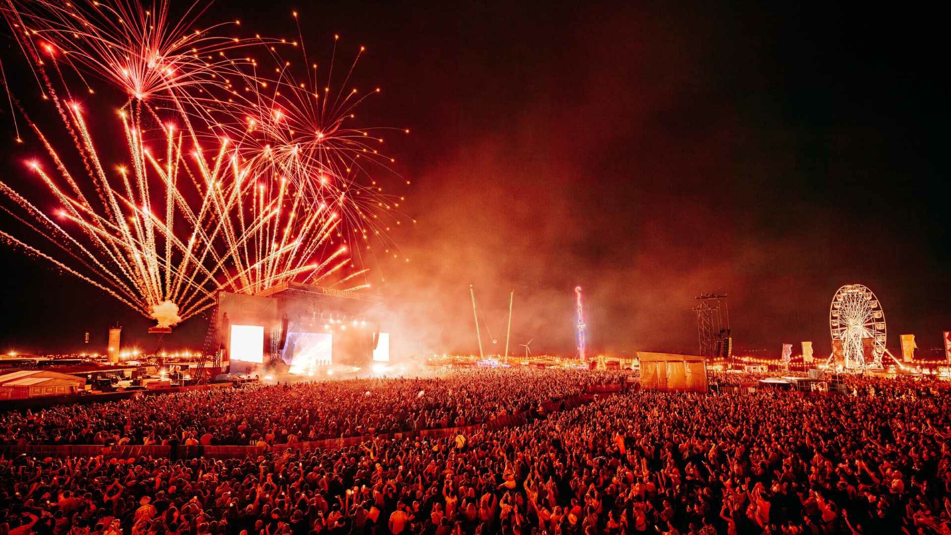 A large crowd at night in front a festival stage lit by red fireworks