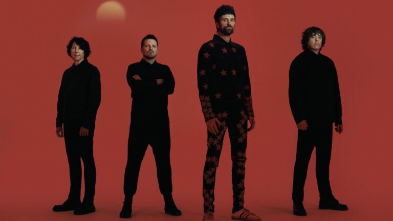 The members of Kasabian standing next to each other wearing black outfits against a red backdrop