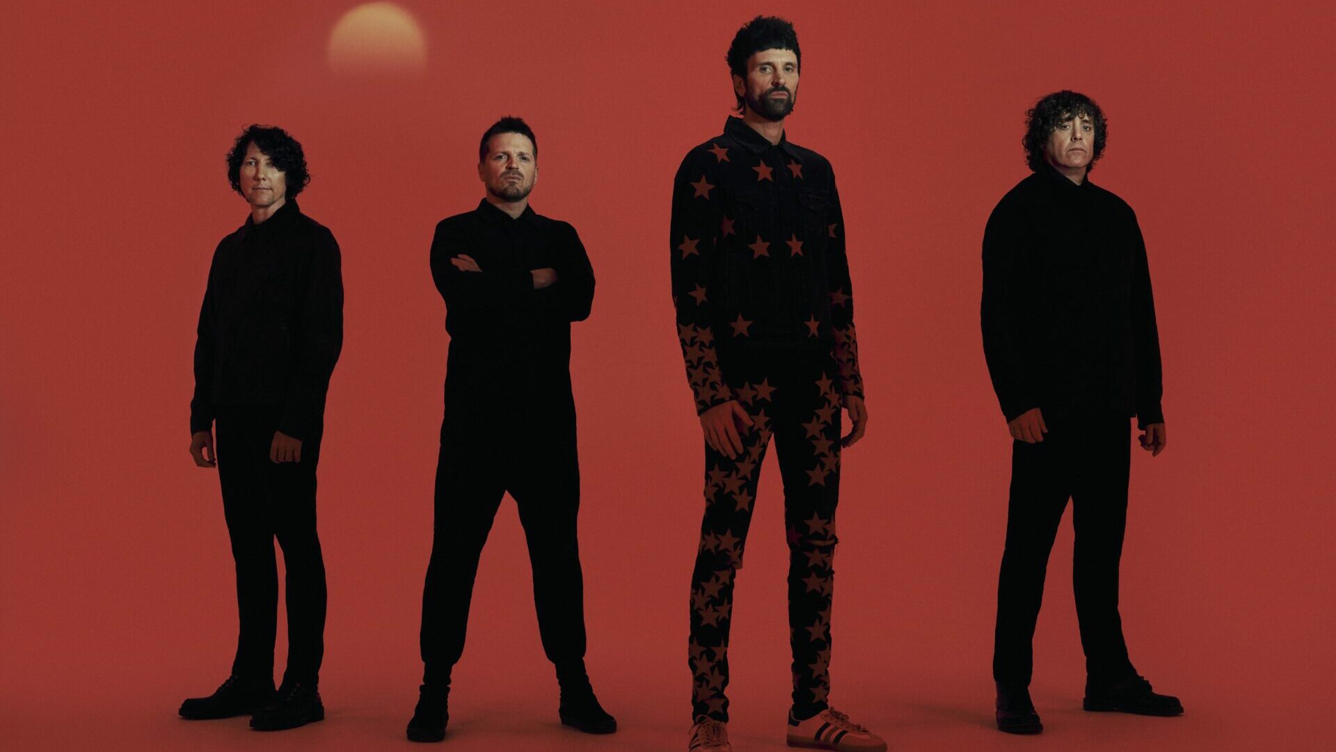 The members of Kasabian standing next to each other wearing black outfits against a red backdrop