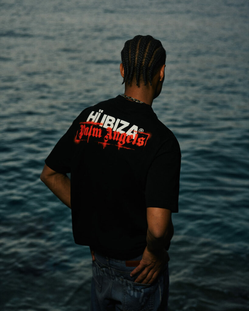 A model wearing a Palm Angel's logo t-shirt with his back turned to the camera facing the ocean. 