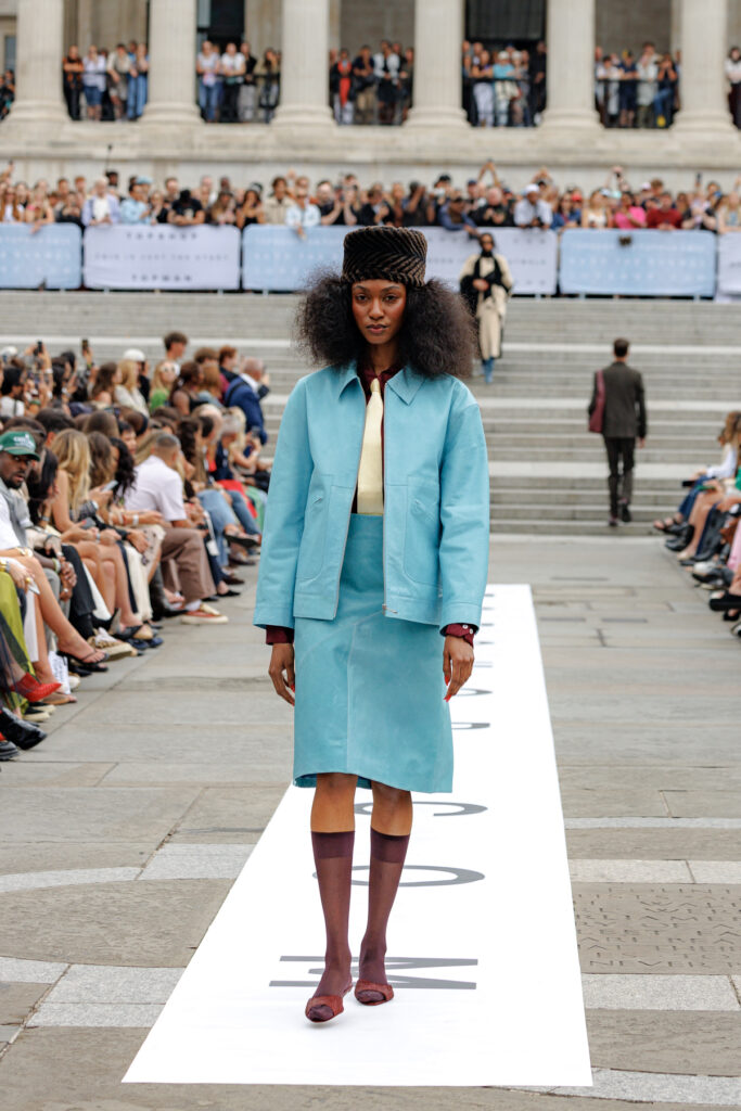 A model wearing a leather turquoise skirt suit, and black pillbox hat walks the Topshop autumn/winter 2025 show in Trafalgar Square.