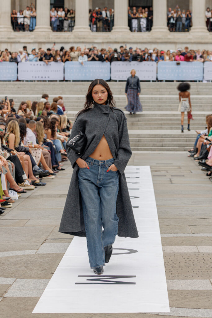 A model wearing a grey trench coat and blue jeans walks the Topshop autumn/winter 2025 show in Trafalgar Square.