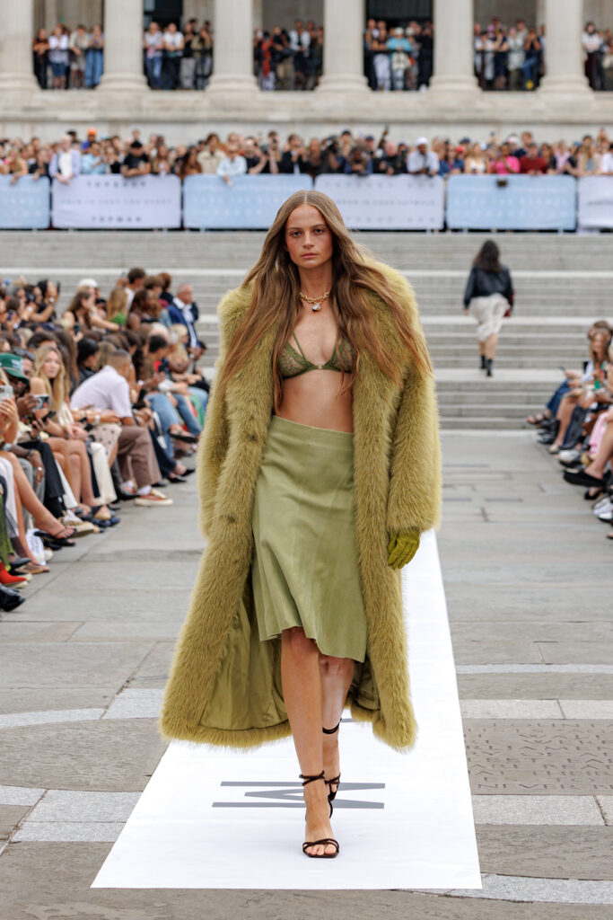 A model wearing a green faux fur coat, lace bra, and green midi length skirt walks the Topshop autumn/winter 2025 show in Trafalgar Square.
