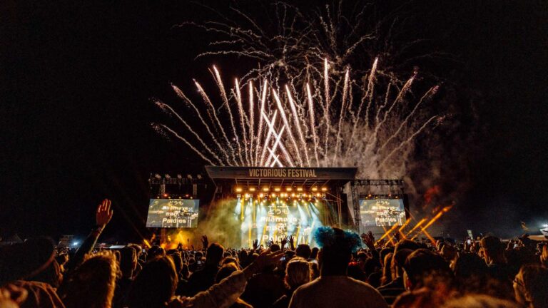 A crowd of people dancing in front of a festival stage with fireworks going off in the background