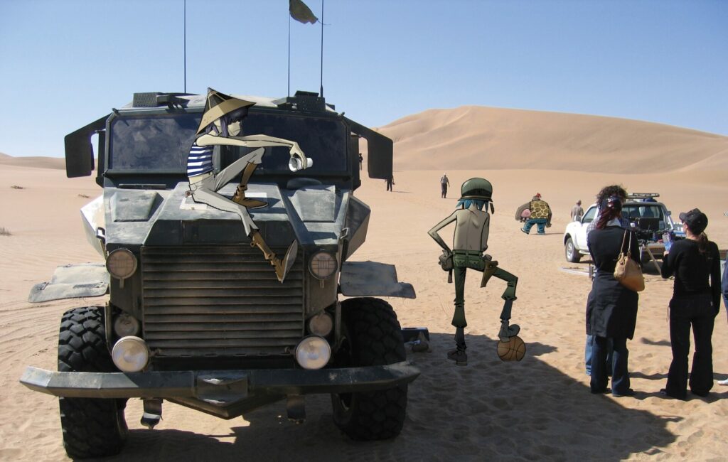 The cartoon members of Gorillaz next to real life people on a video shoot in the desert