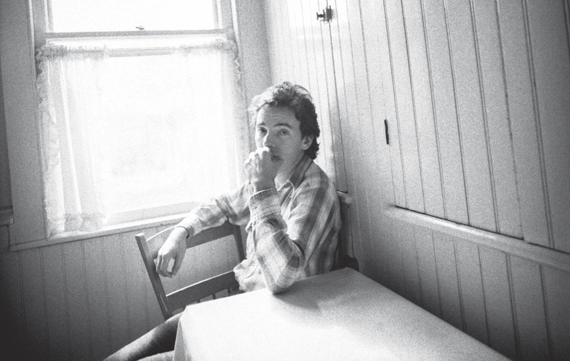 Bruce Springsteen in black-and-white sat at a kitchen table by a window