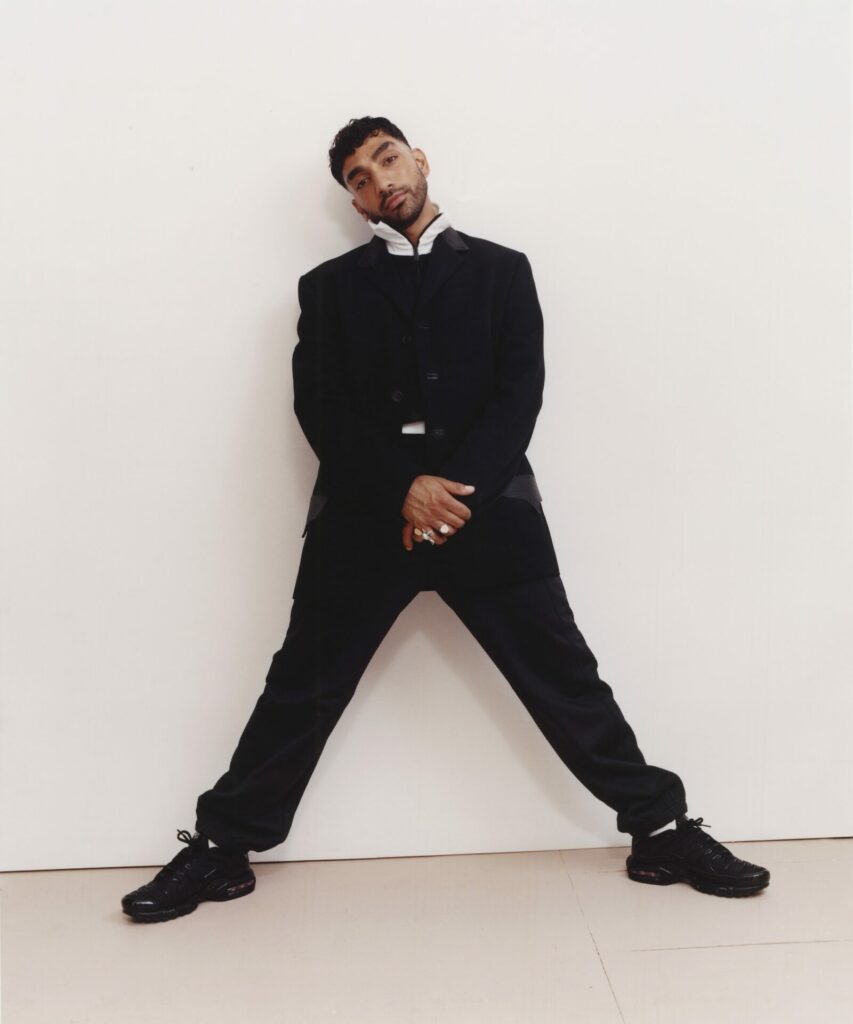 Mawaan Rizwan posing for a photoshoot with his legs apart and leaning against a wall