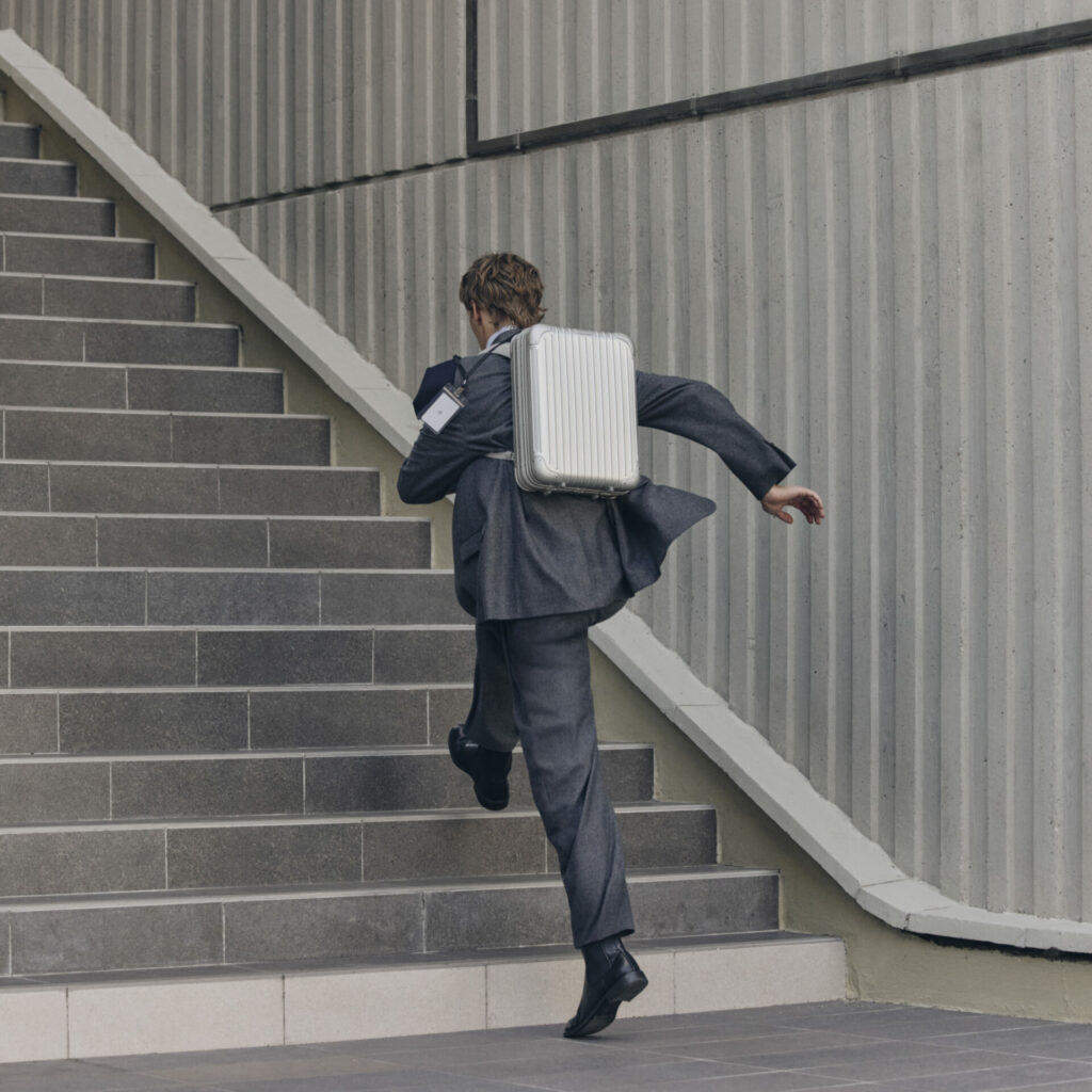 Lifestyle shot of the RIMOWA Original backpack. A model in a suit jumps up the stairs carrying an aluminium backpack.