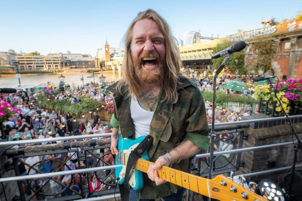 Sam Ryder holding a guitar and laughing looking into the camera with a large audience behind and below him as he stands on a balcony of a pub with Big Ben visible in the background
