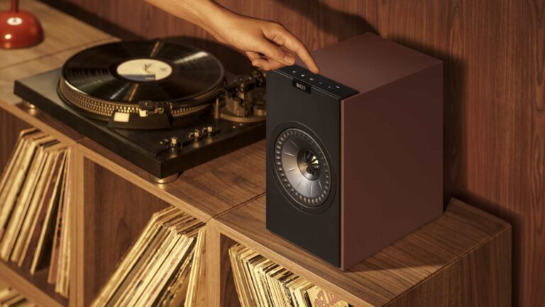A KEF Coda W speaker in Vintage Burgundy, positioned beside a turntable on a wooden shelf unit stacked with vinyl records.